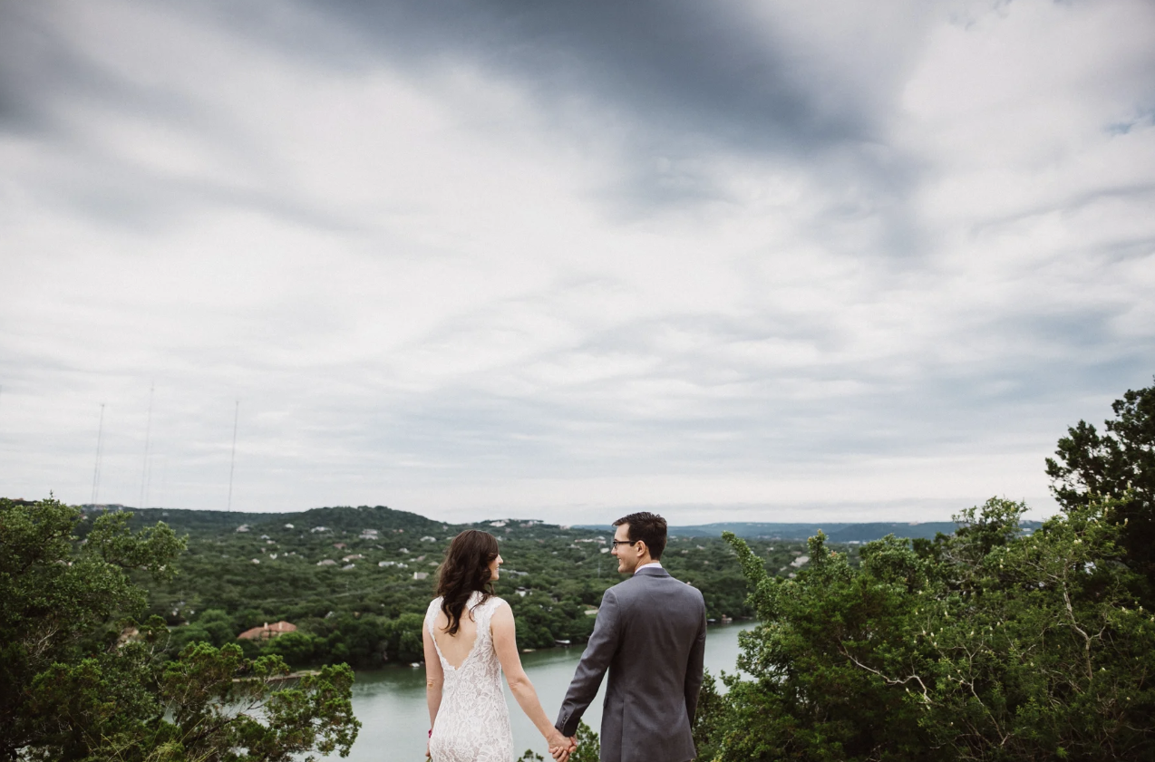 A couple holding hands and facing each other outdoors, with a view of a river, greenery, and a hilly landscape under a cloudy sky.