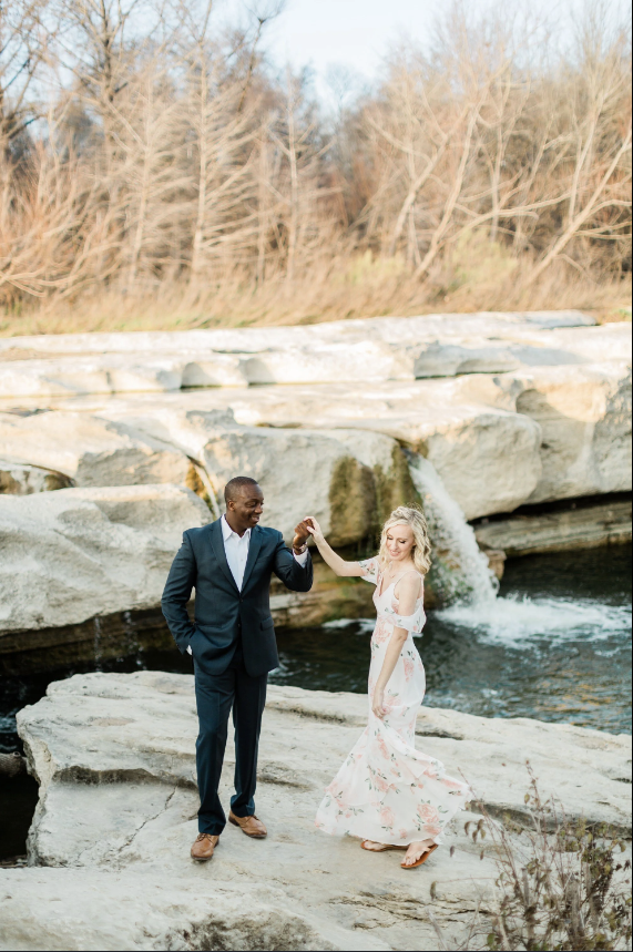 A couple dancing on rocks by a river, with a waterfall in the background, during daytime.