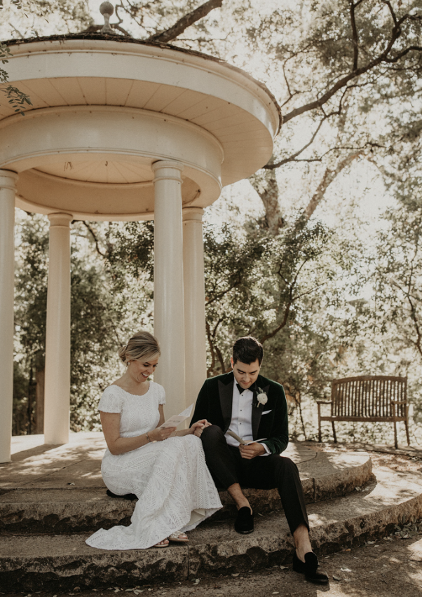 A bride and groom sitting on the steps of a white pavilion outdoors, reading something together, surrounded by trees.