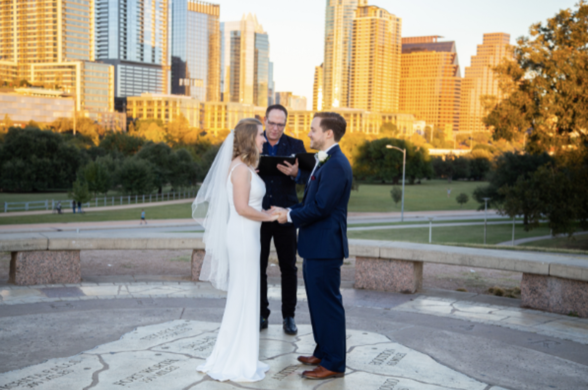 A couple gets married outdoors in a city park during sunset, with skyscrapers in the background. The bride wears a white gown and veil, and the groom is in a blue suit. They hold hands and look at each other as an officiant conducts the ceremony.