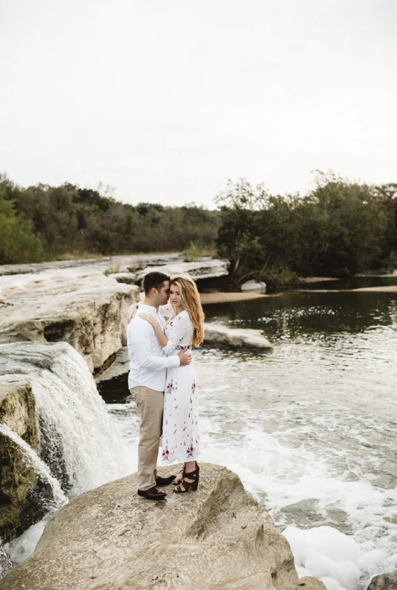 A couple stands on a large rock next to a waterfall and river with trees in the background, sharing an intimate moment.