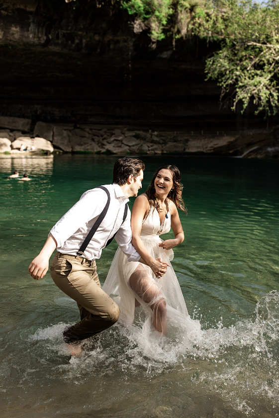 A couple in wedding attire playing and laughing in a river surrounded by rocky cliffs and green trees.