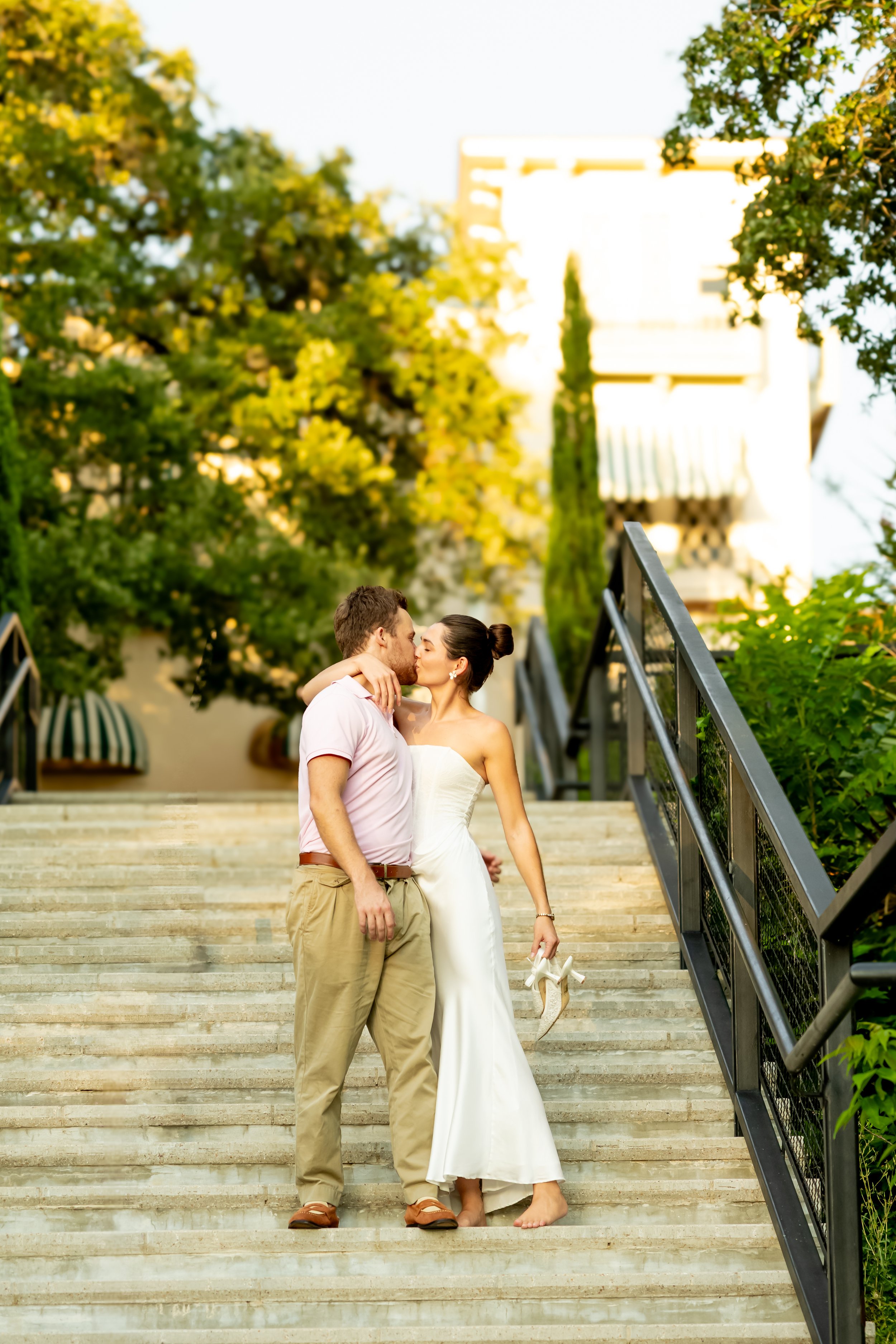 A couple sharing a kiss on outdoors steps in warm sunlight, with lush trees and a white building in the background.