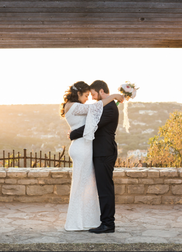 Bride and groom embrace under a wooden roof with a scenic countryside background at sunset.