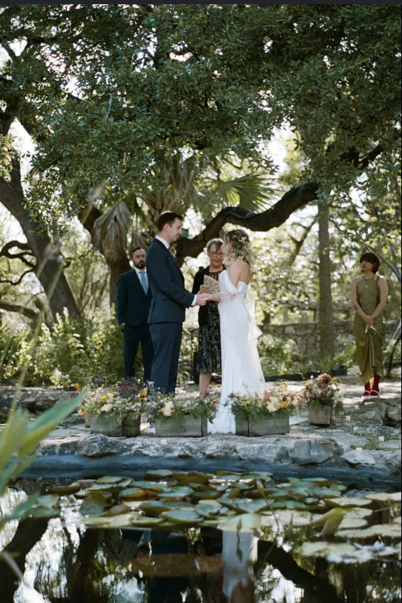 A couple getting married outdoors during the day, standing before an officiant, with a pond in the foreground and trees in the background.