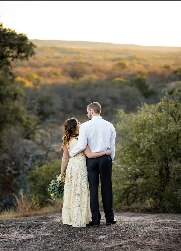 A couple embracing outdoors on a rocky platform with a scenic backdrop of hills and trees during sunset, with the woman holding a bouquet.