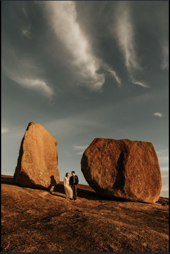 A couple dressed in formal attire standing between two large, weathered rocks on a hillside during sunset, with a dramatic sky and clouds overhead.