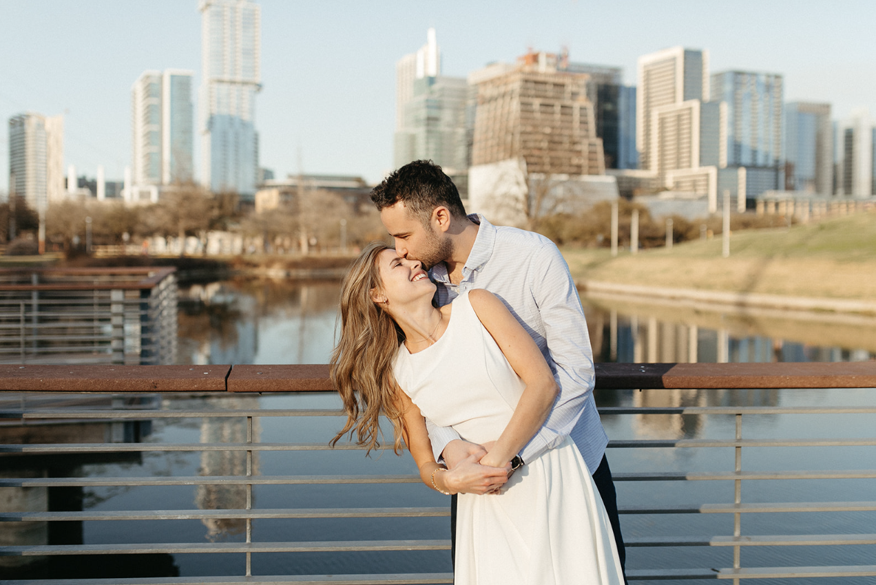 A couple embraces on a bridge over a body of water with a city skyline in the background.