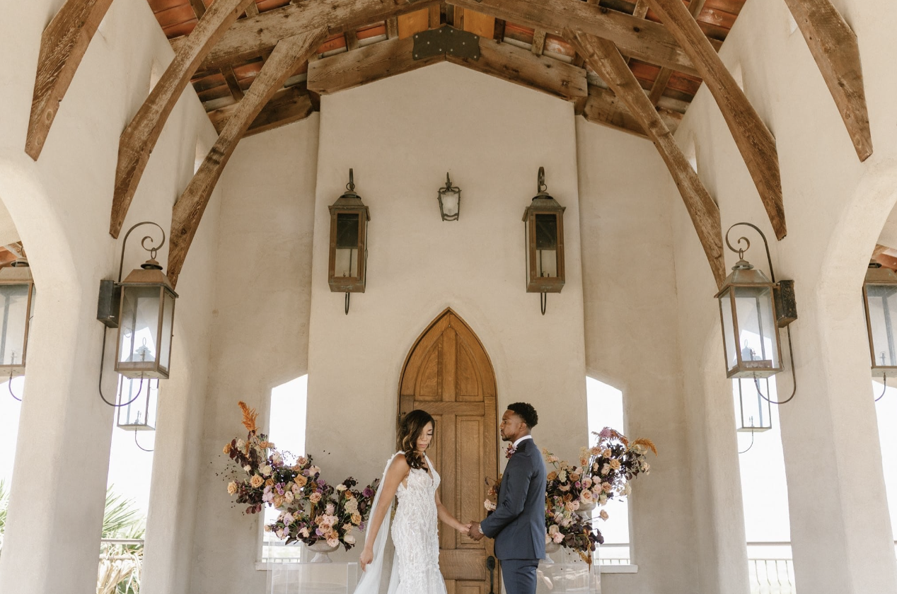 A bride and groom holding hands in a wedding ceremony inside a chapel with white walls, wooden beams, and large floral arrangements on either side.