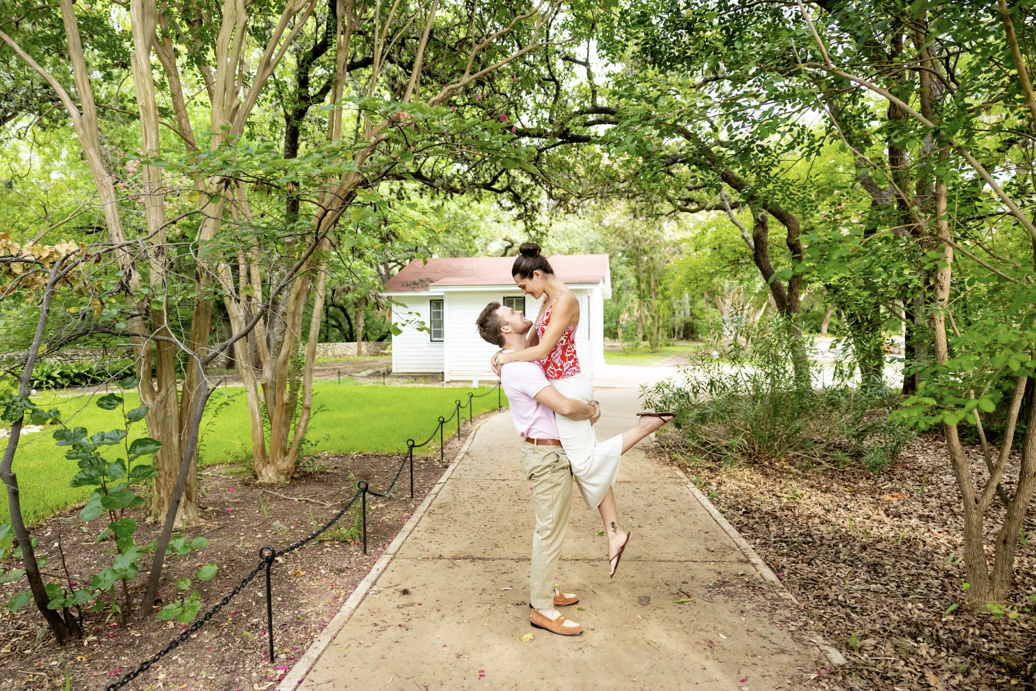 A happy couple is outdoors on a garden path, with the man lifting the woman in his arms. They are smiling at each other affectionately. The man is wearing tan pants and a light pink shirt, while the woman is wearing a red and white patterned top and white pants. There is greenery and trees around them, with a small white house visible in the background.