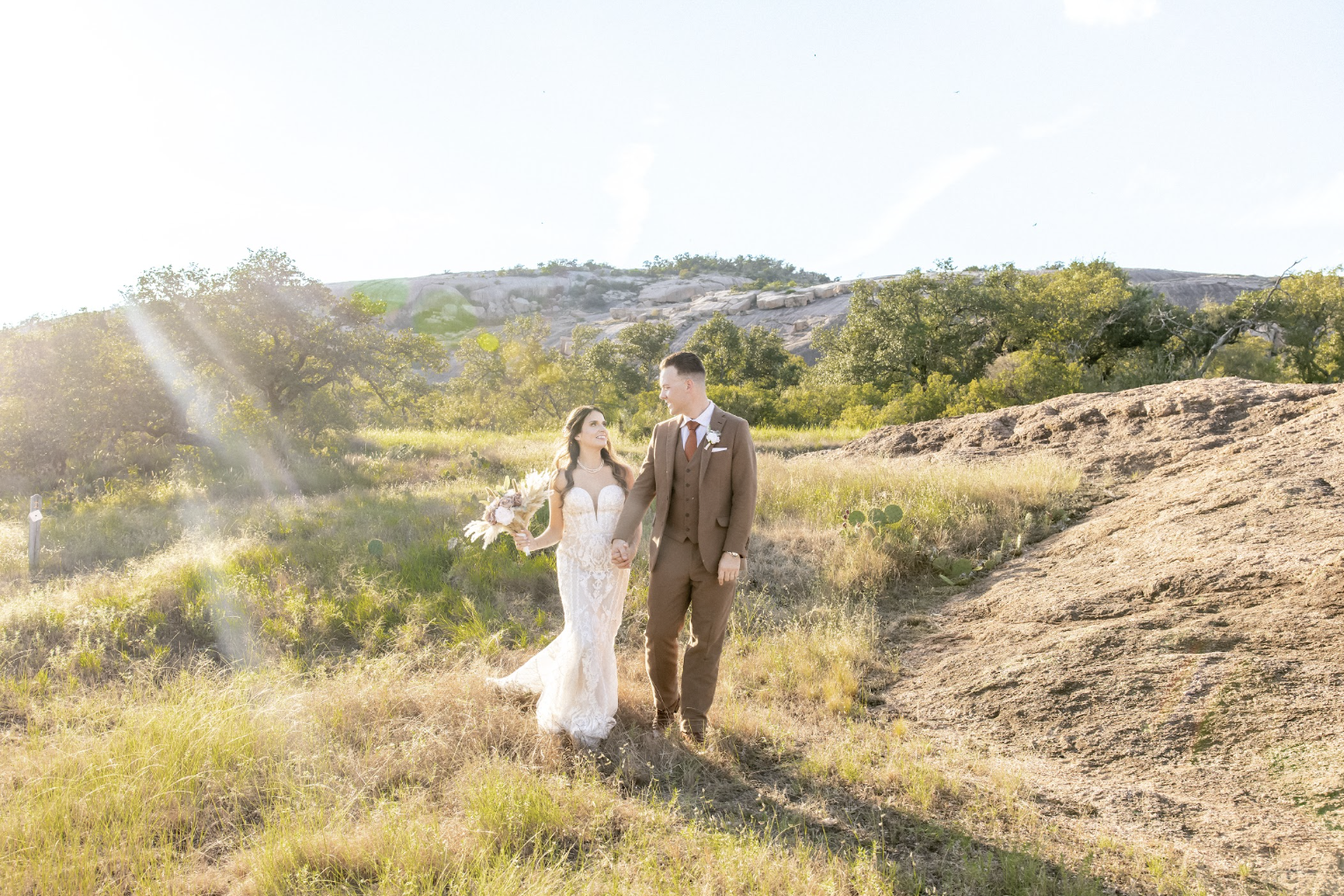 A bride and groom holding hands and walking outdoors on a sunny day, with trees and rocks in the background.