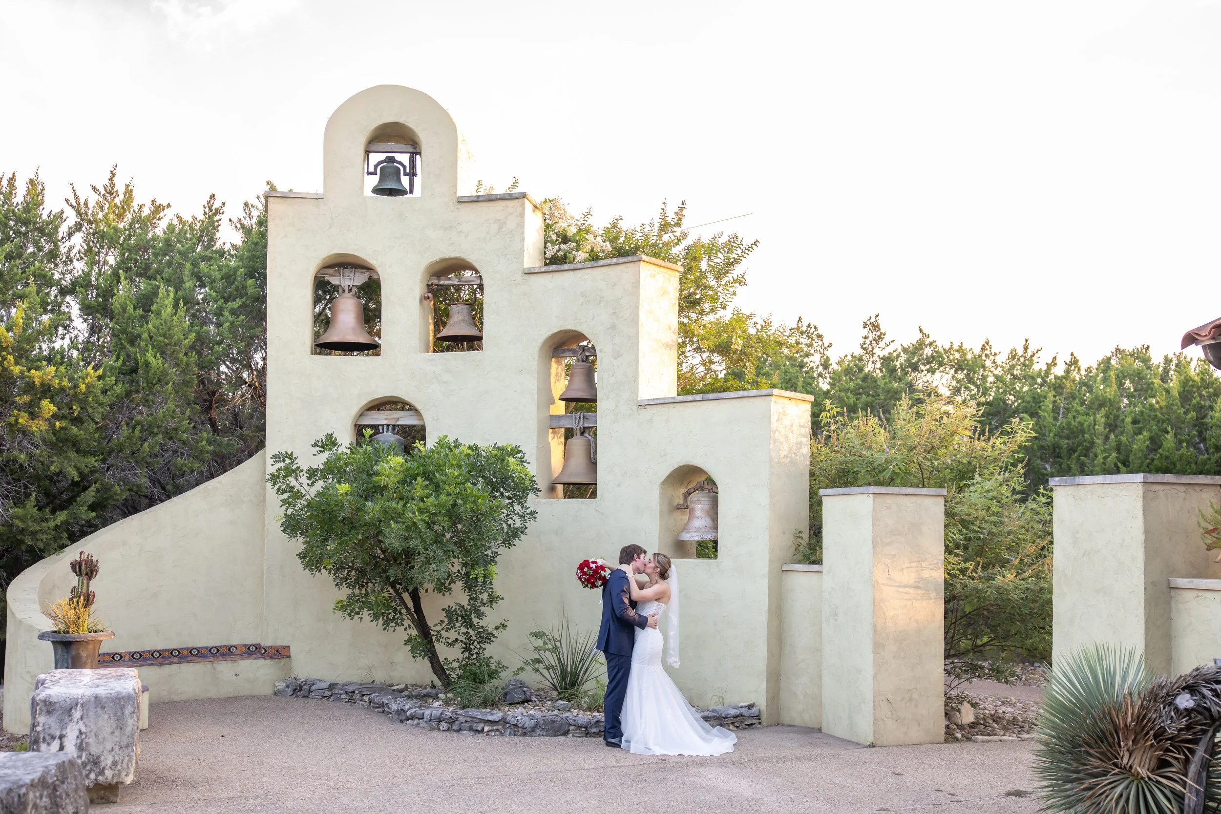 A couple in wedding attire sharing a kiss in front of a Spanish-style bell tower with greenery and trees in the background.
