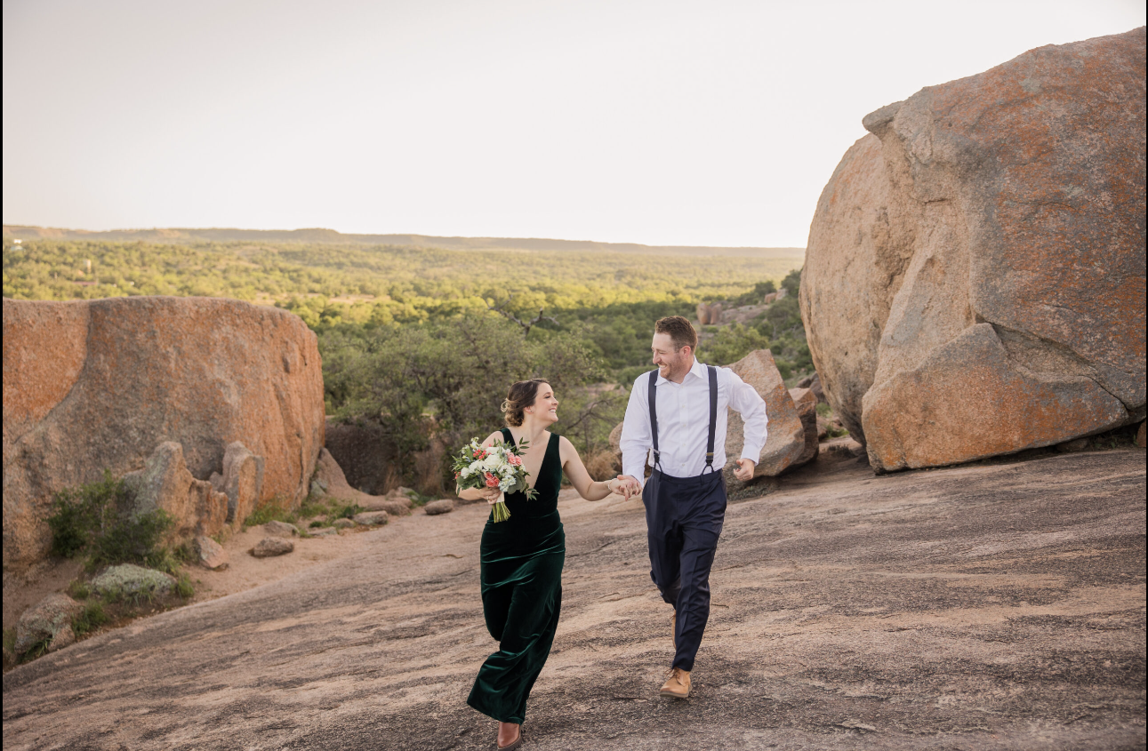 A couple is running hand in hand on rocky terrain outdoors at sunset, with large boulders and green trees in the background. The woman is holding a bouquet of flowers and is wearing a dark green velvet dress, while the man is dressed in a white shirt with suspenders and dark pants.
