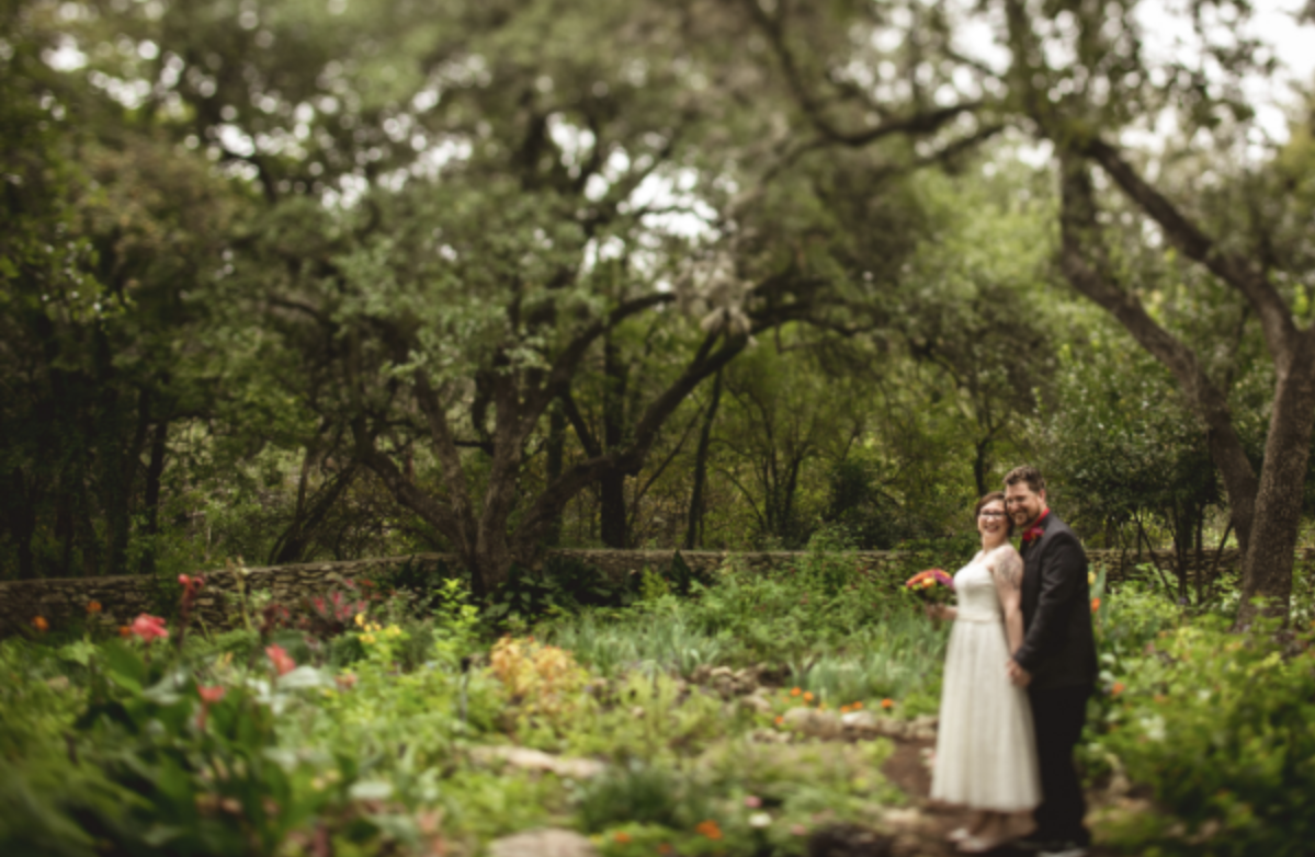 A couple in wedding attire standing in a lush garden with trees and flowers, smiling at the camera.