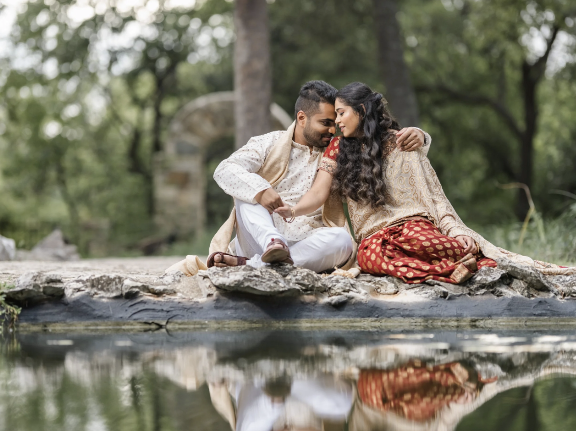 A couple dressed in traditional Indian clothing sitting on a rock by a river, lovingly looking at each other.