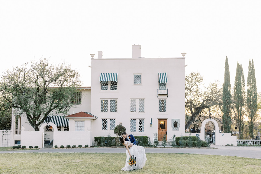A couple dancing outside a white multi-story house with green trees and bushes, during sunset.