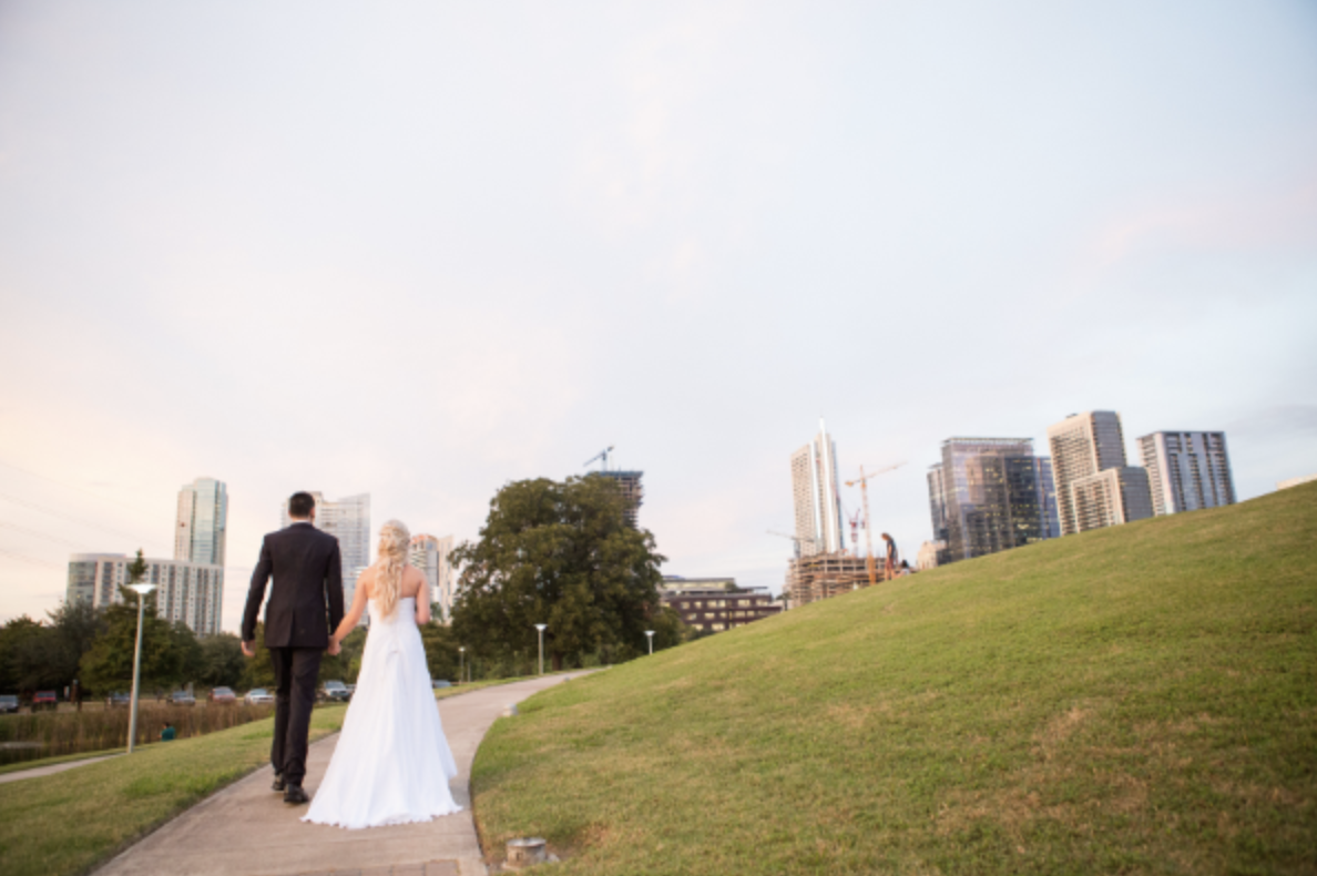 A newlywed couple holding hands, walking on a park pathway with a city skyline in the background during sunset.