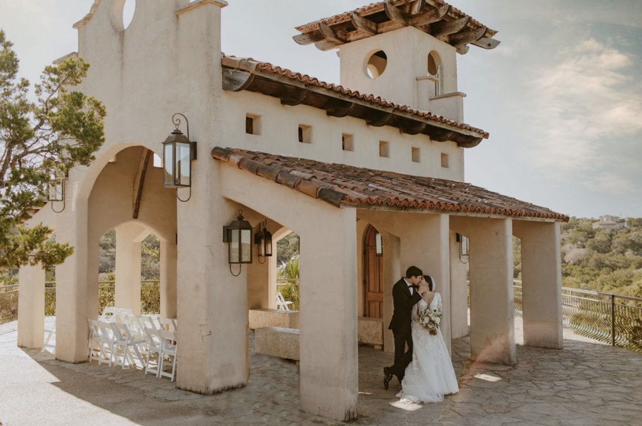 Bride and groom sharing a kiss outside a small beige stucco chapel with a red tile roof, surrounded by trees and a scenic view.