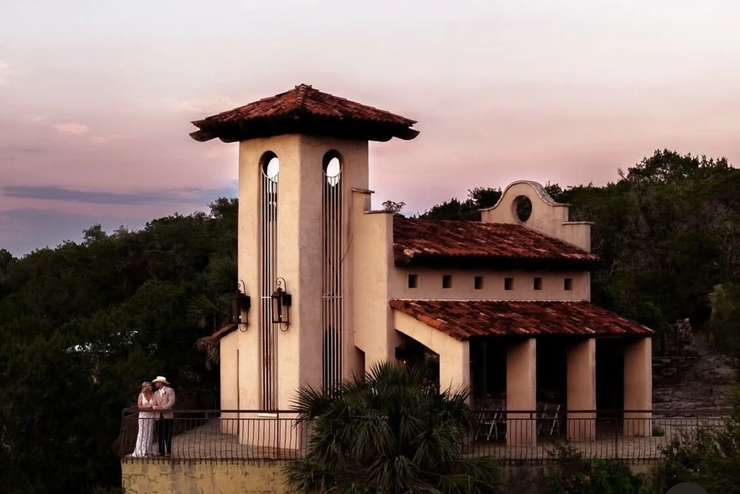 A small Mediterranean-style building with a tower, red tile roof, surrounded by greenery at sunset, with a couple standing on a balcony.