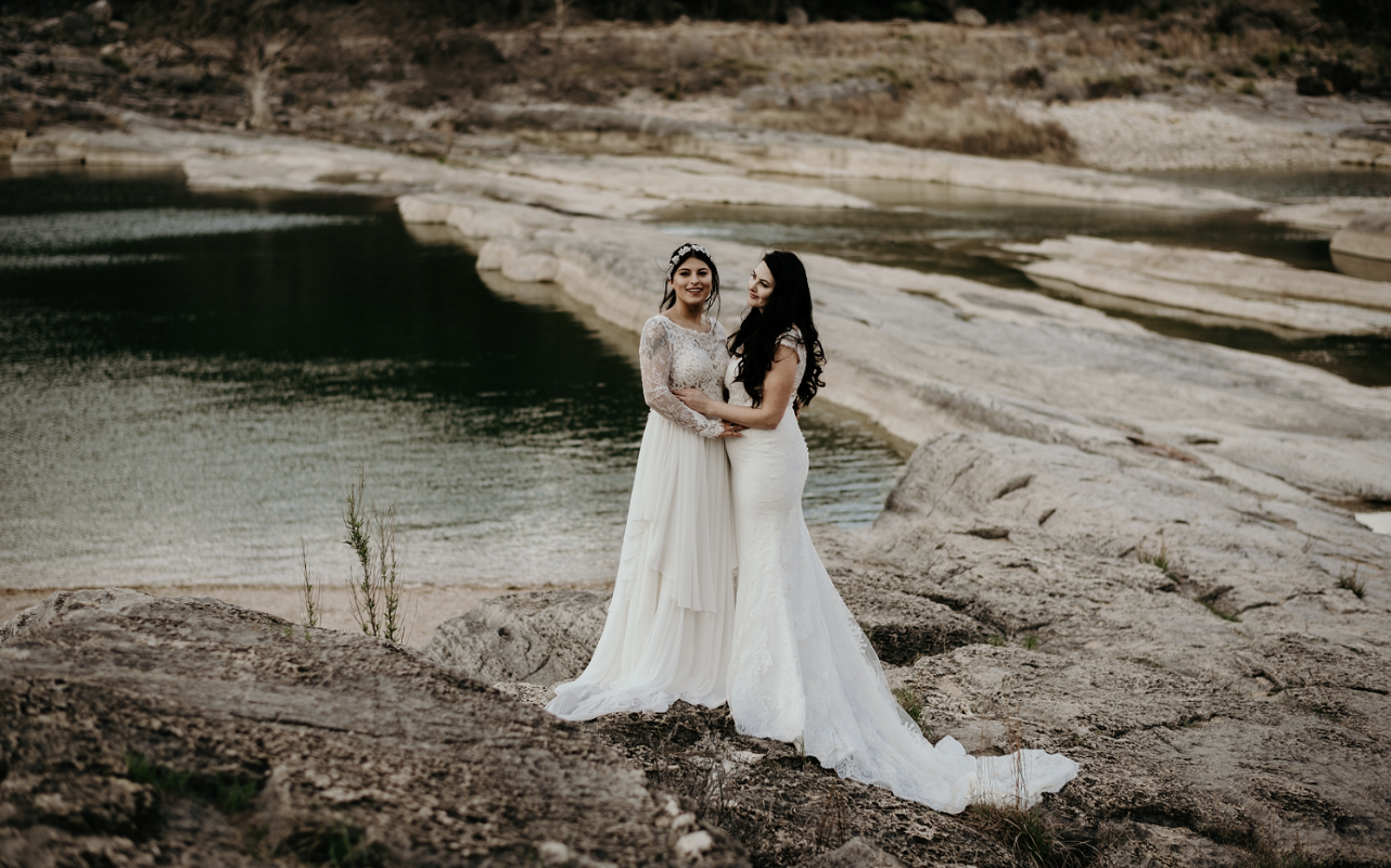 Two women in wedding dresses standing on rocks by a body of water, smiling and holding hands, in an outdoor natural setting.