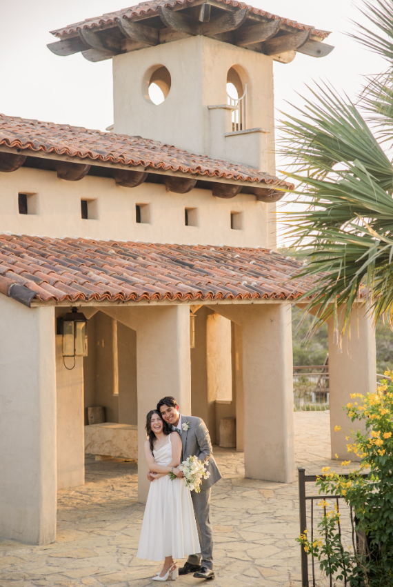 A happy couple in wedding attire standing in front of a Mediterranean-style building with stucco walls and a tiled roof, during sunset.
