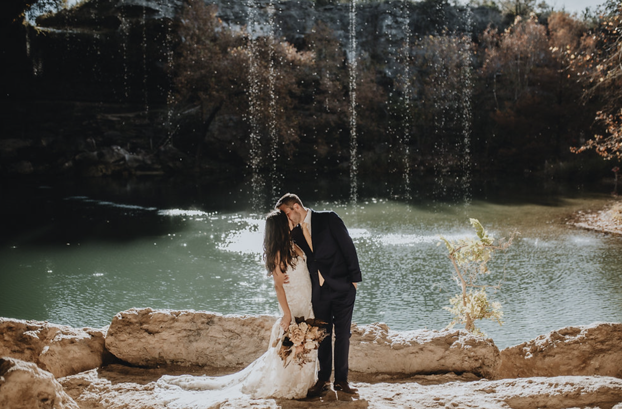 A bride and groom kissing by a waterfall behind a lake surrounded by autumn trees.