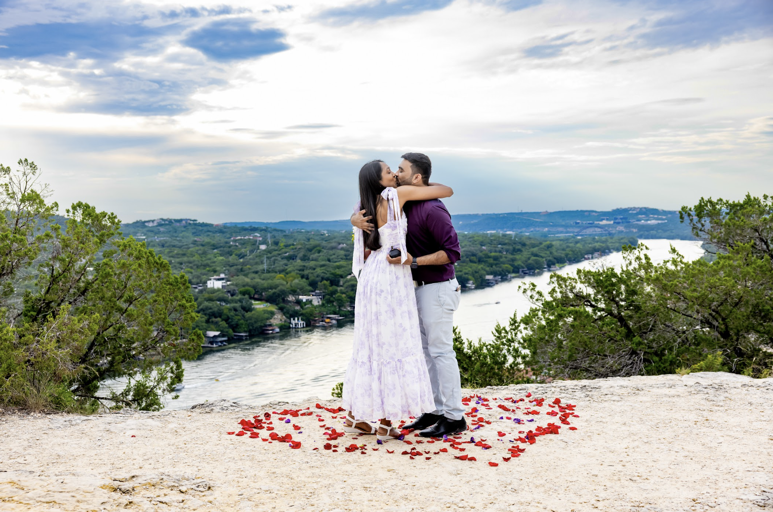 A couple hugging and kissing on a scenic overlook with a river, green trees, and a cloudy sky in the background, surrounded by red flower petals on the ground.