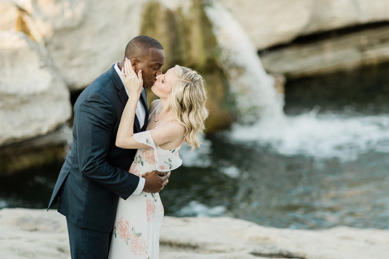 A couple sharing a romantic kiss by a river with rocks and flowing water in the background.