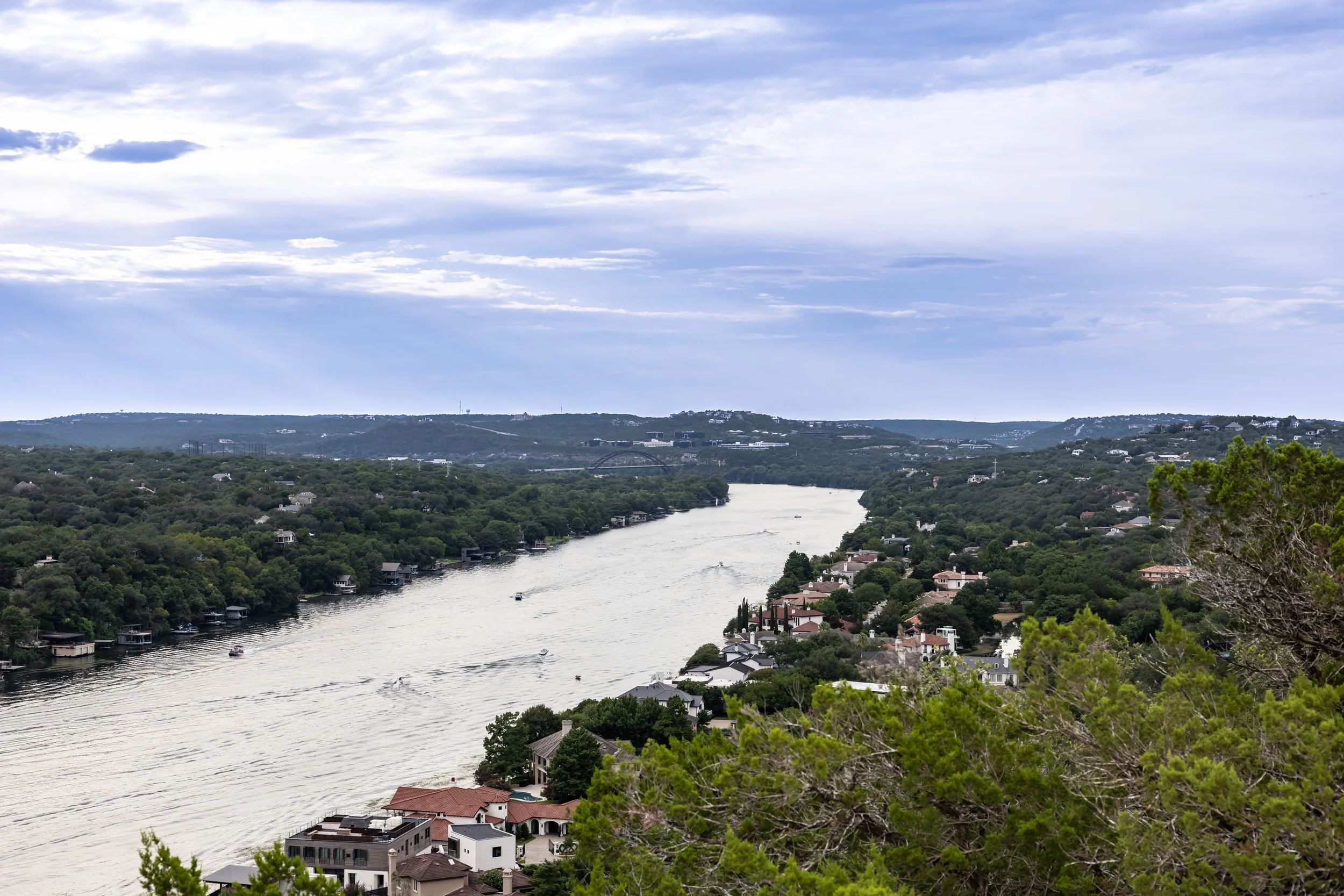 A scenic view of a river flowing through a lush, green landscape with houses along the shoreline and across the water. The sky is partly cloudy with patches of blue.