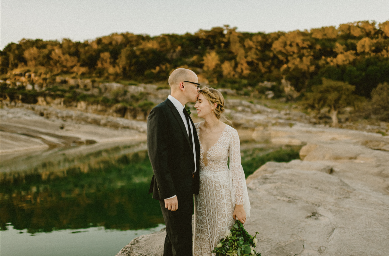 A newlywed couple in formal wedding attire standing on a rocky outcrop by a lake, with a backdrop of trees and hills during golden hour.