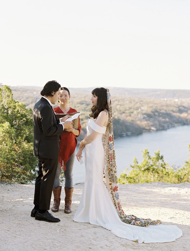 A couple gets married outdoors by a lake with two officiants present, one reading from a book, as the bride and groom look at each other.