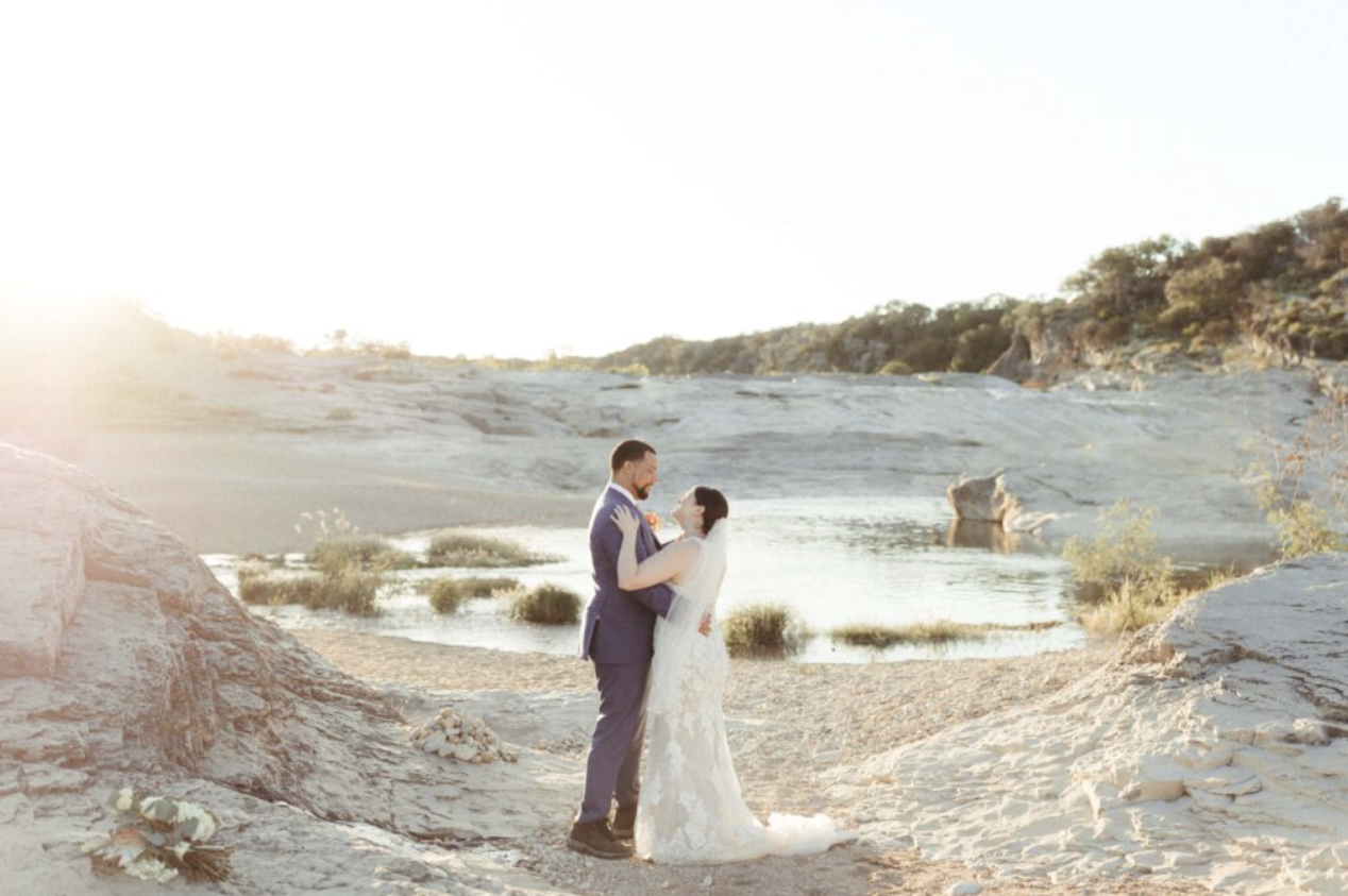 A bride and groom in wedding attire holding each other and smiling by a river at sunset with a rocky landscape in the background.