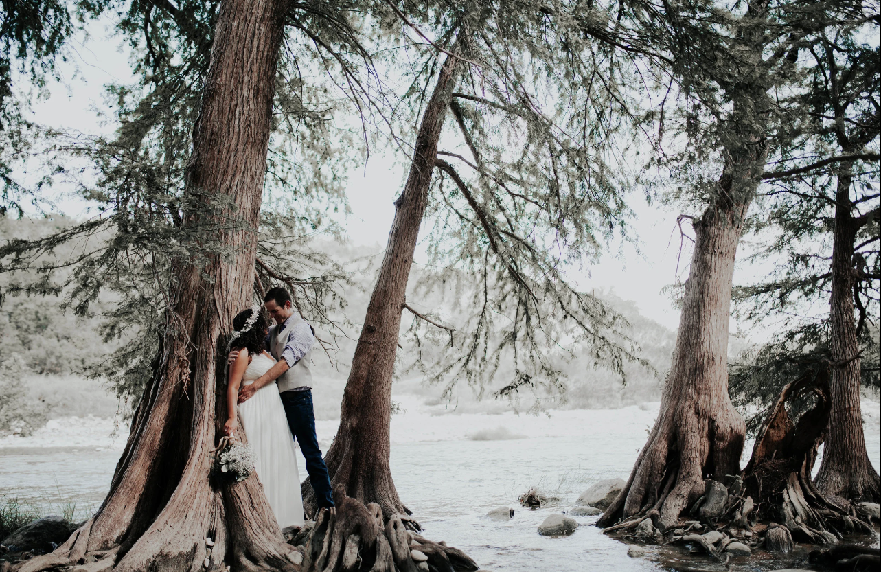A couple dressed in wedding attire standing by a large tree and holding hands near a river. The woman is holding a bouquet, and the scene is outdoors with trees and water in the background.