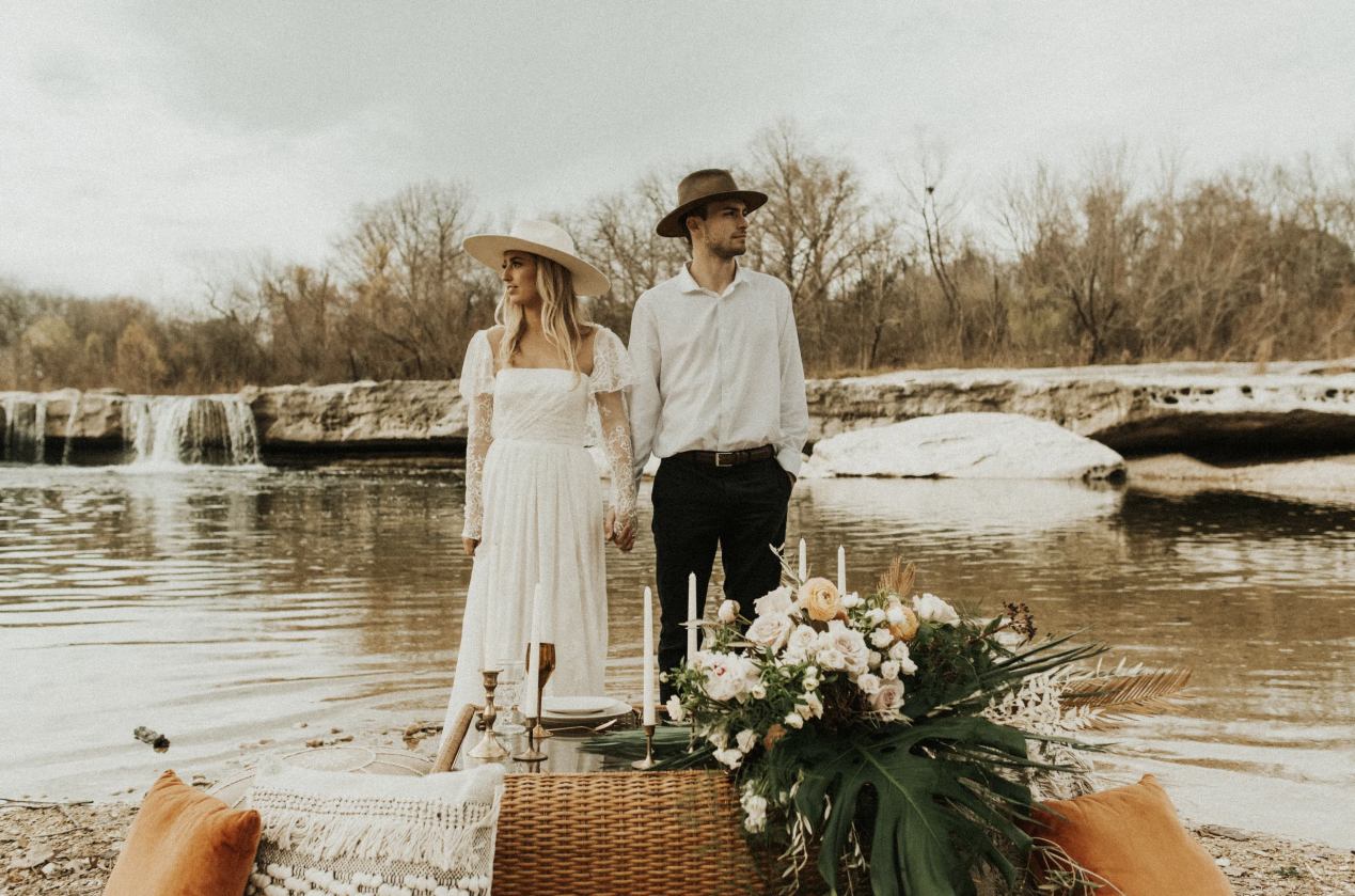 A couple stands by a river with a waterfall in the background, dressed in vintage bohemian wedding attire, holding hands, with a decorated table and floral arrangement in the foreground.