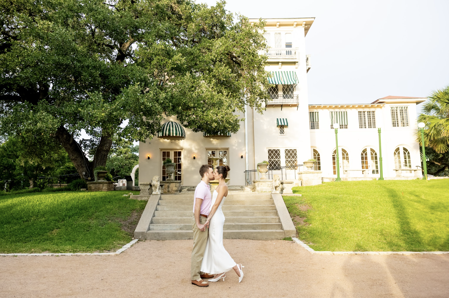 A couple kissing in front of a large white house with green awnings, surrounded by lush green trees and grass.