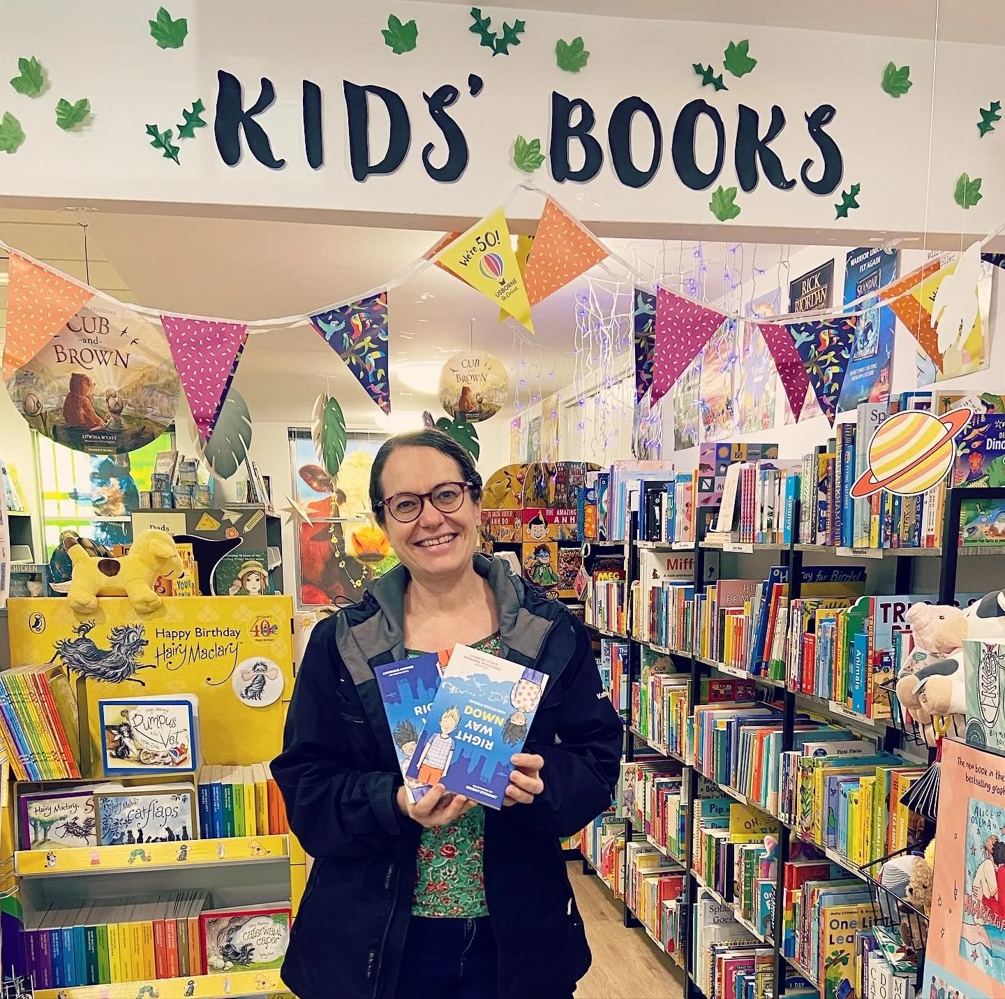 Rebecca stands in a bookshop under a sign that says Kids' Books. She is surrounded by shelves full of books. 
