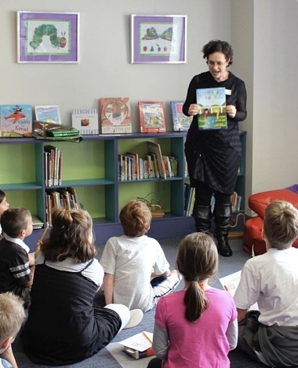 Woman in library holds book up to show group of children sitting at her feet.