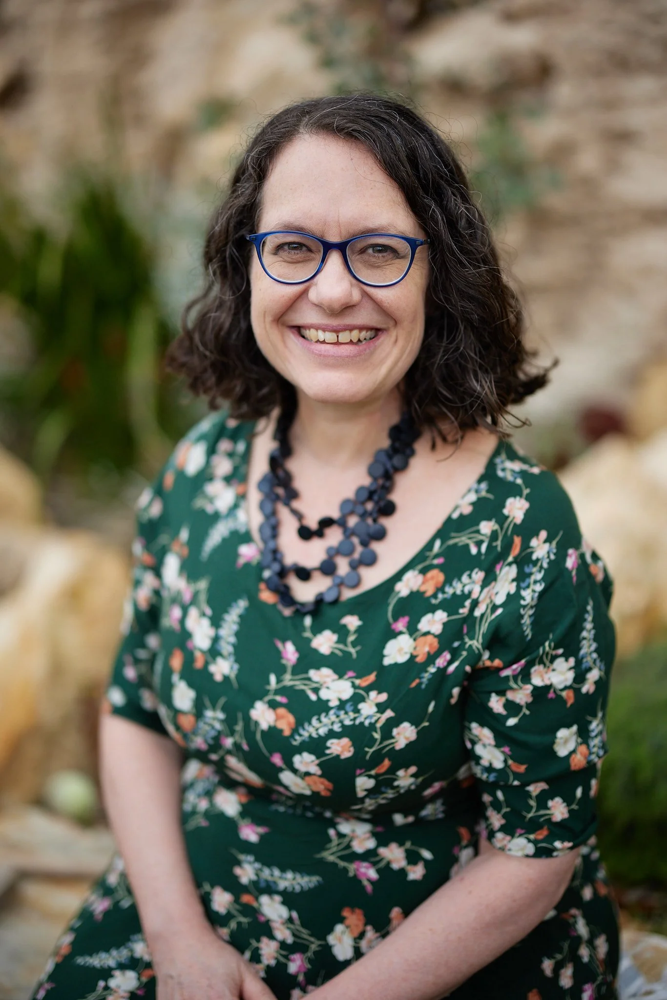 Rebecca head and shoulders photo. Rebecca has short brown hair, blue-framed glasses, green floral dress and black beaded necklace.
