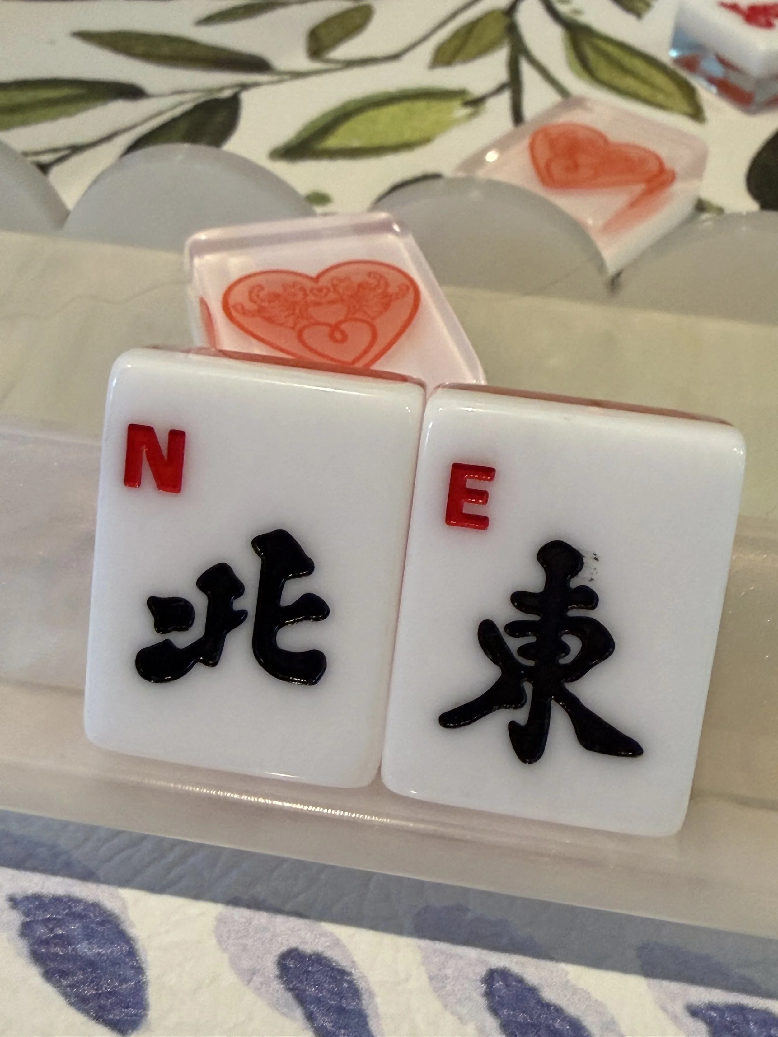 Close-up of white tiles with black Chinese characters, some with red letters, on a table with a floral tablecloth.