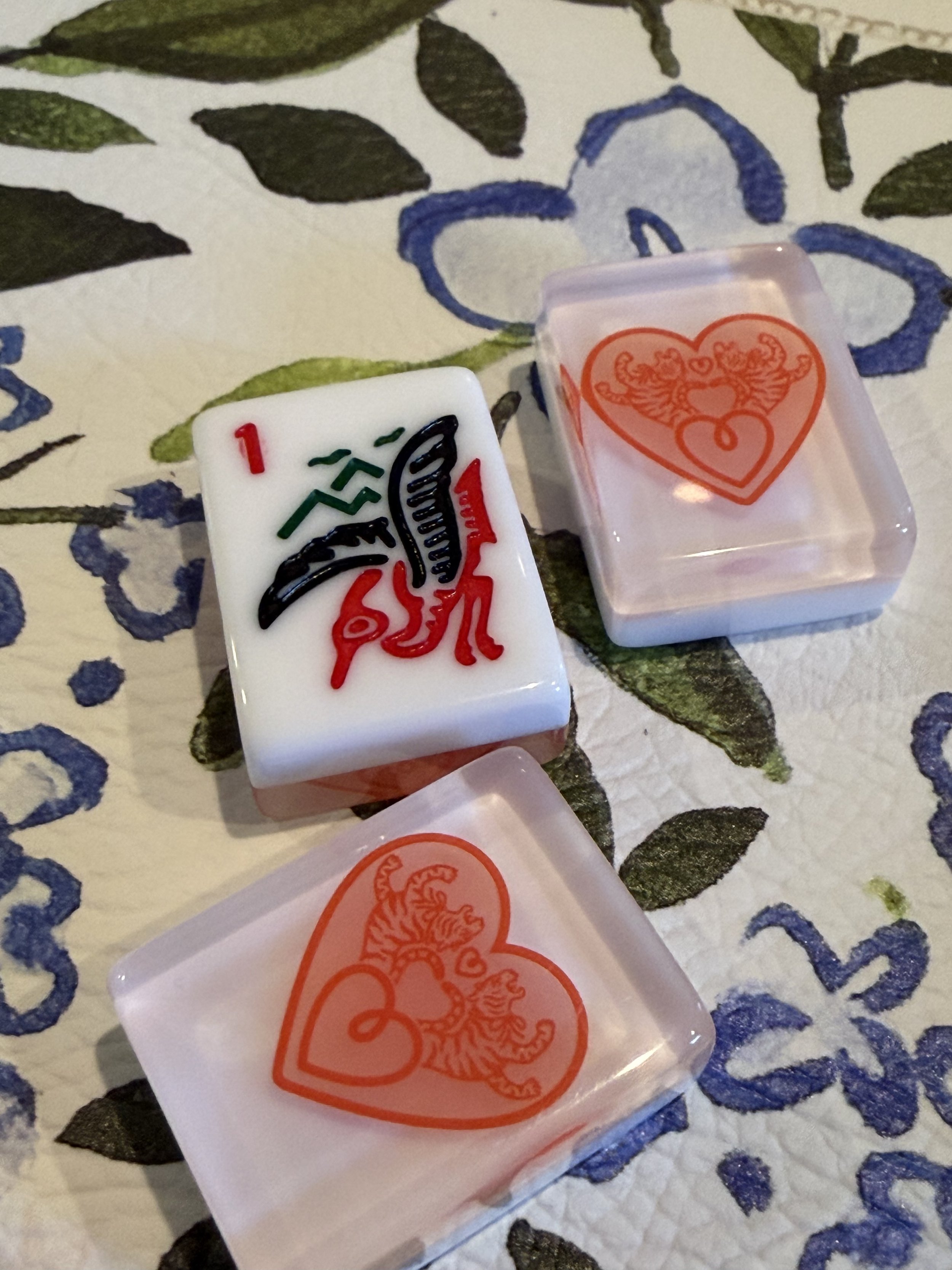 Close-up of two plastic objects resting on a textured fabric surface with a floral pattern. One object is white with black, green, and red designs, while the other is clear with an orange tiger illustration inside a heart shape.
