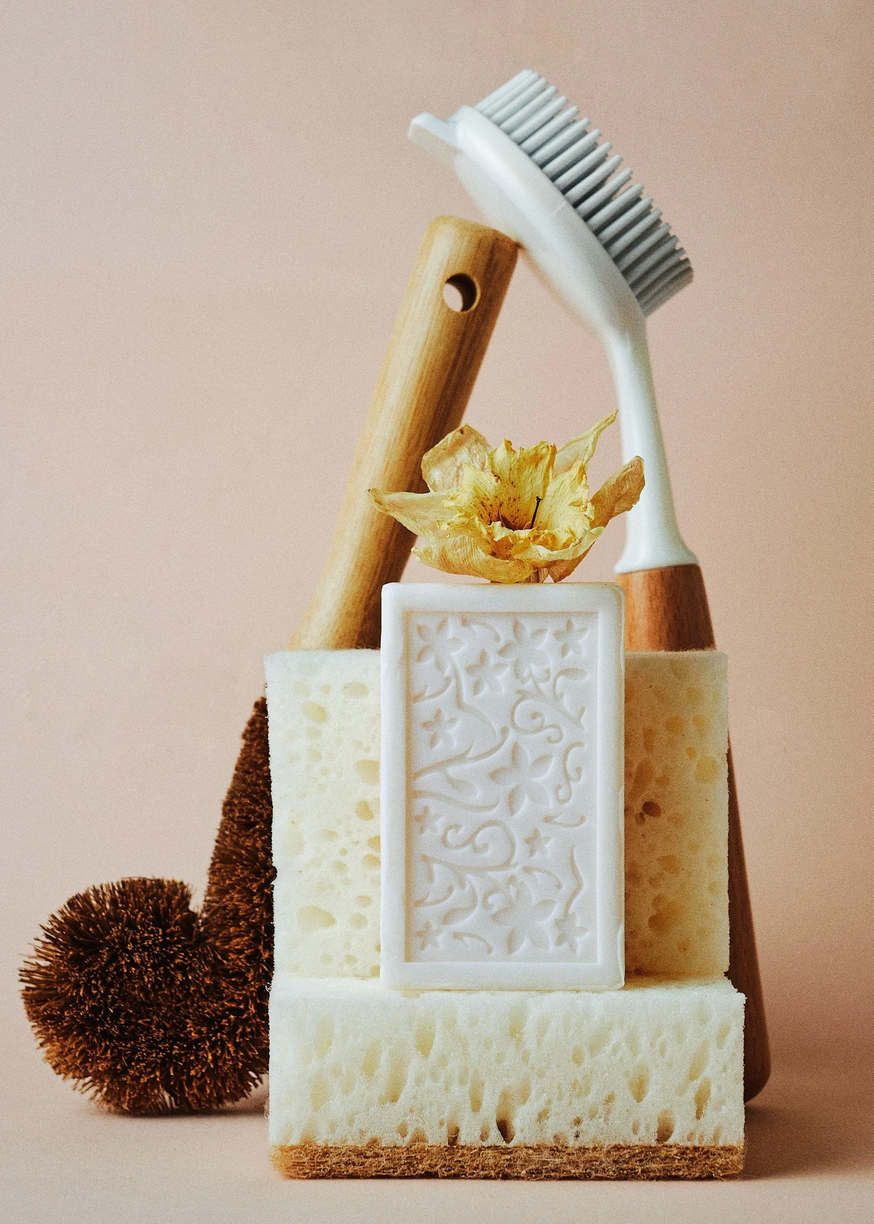 A composition of various cheeses, a wooden-handled brush, a dried flower, a brown textured sponge, and a soap with intricate patterns, arranged vertically against a neutral background.