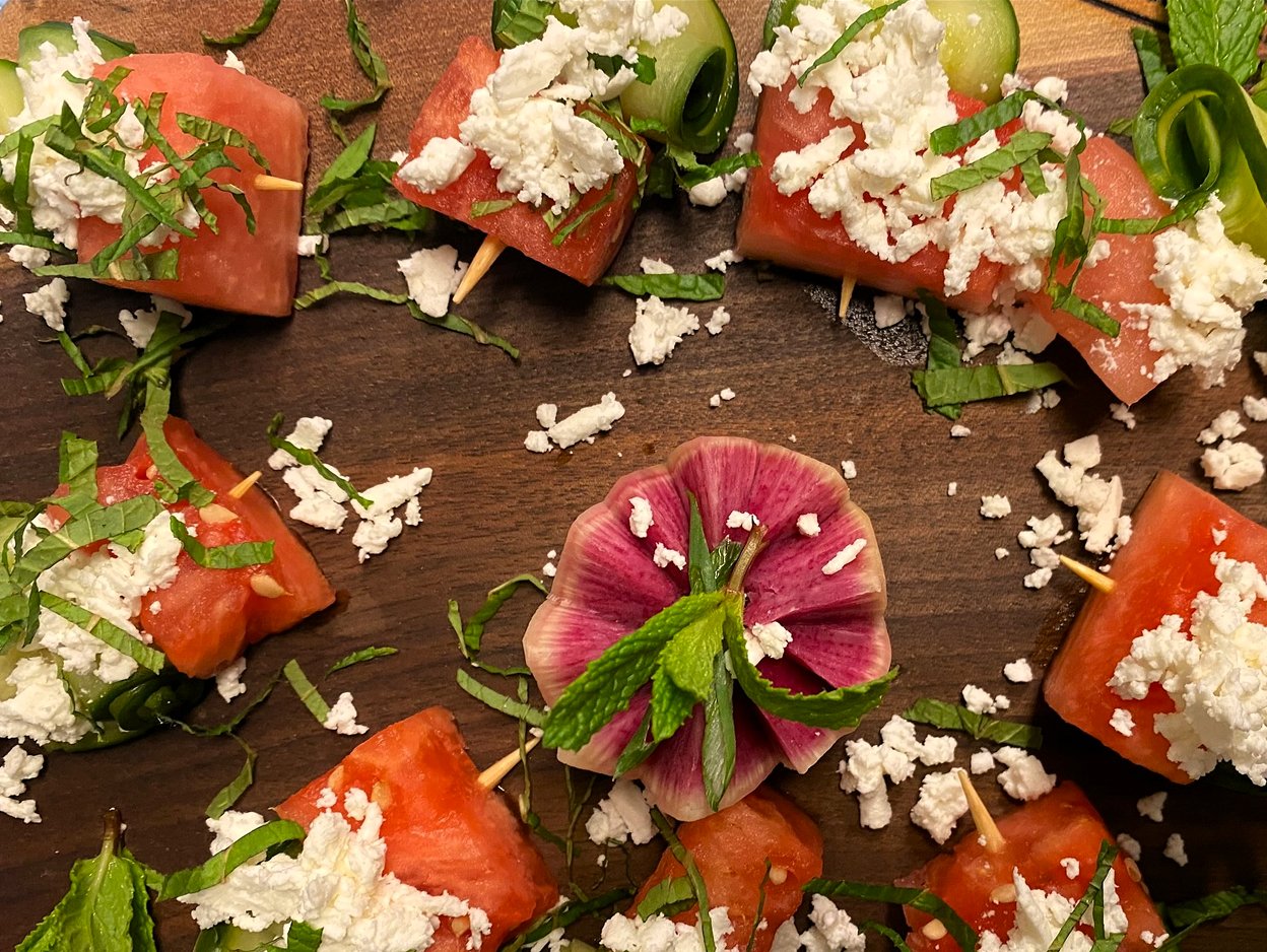 Arranged Hors D'oeuvres with watermelon cubes topped with crumbled feta, chopped green herbs, and cucumber slices on a wooden surface.