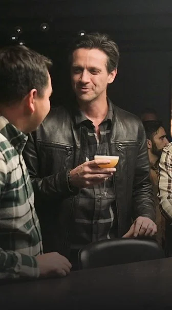 A man in a black leather jacket holding a cocktail while conversing with another man in a striped shirt at a social gathering.
