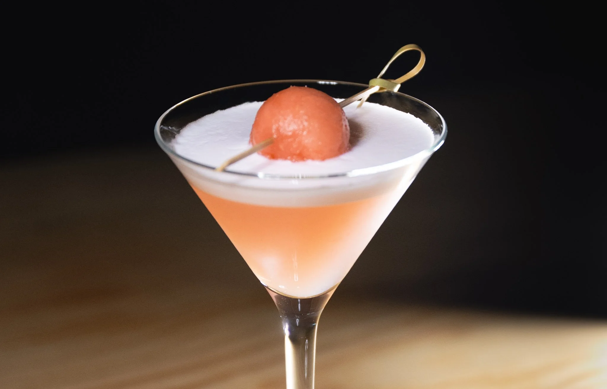 Sandìa in a martini glass topped with foam, garnished with a watermelon ball on a pick, against a dark background.