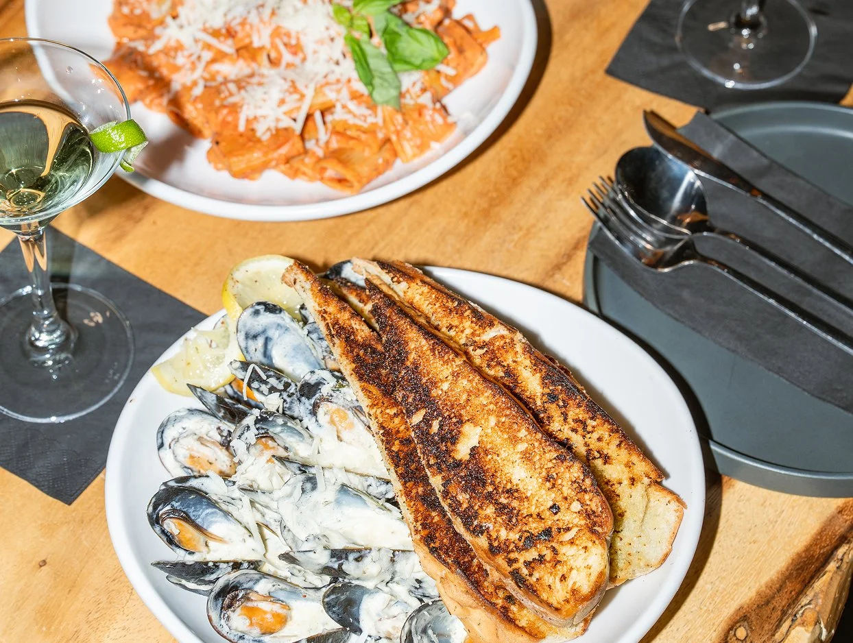Restaurant meal featuring mussels in a creamy sauce with focaccia, pasta topped with tomato sauce and grated cheese, and refreshing beverages on a wooden table.
