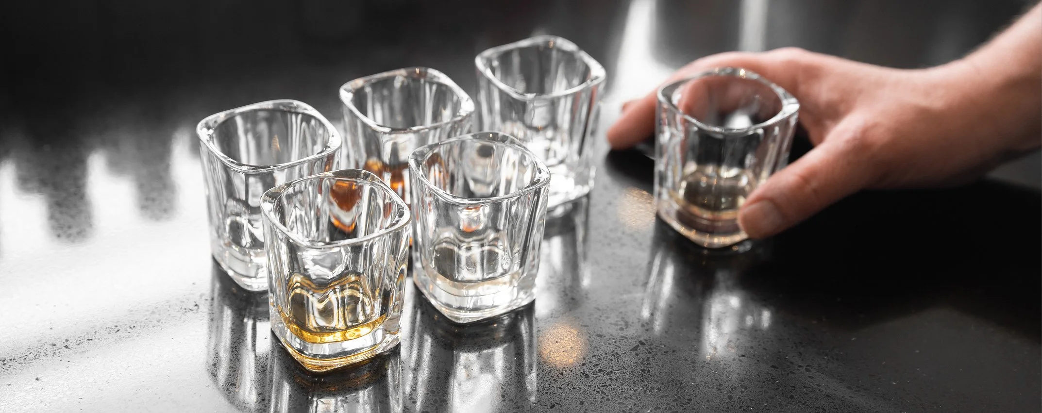 Several square-shaped tasting glasses containing a variety of spirits and liqueurs, placed on a shinny black countertop. A person is grabbing a glass.