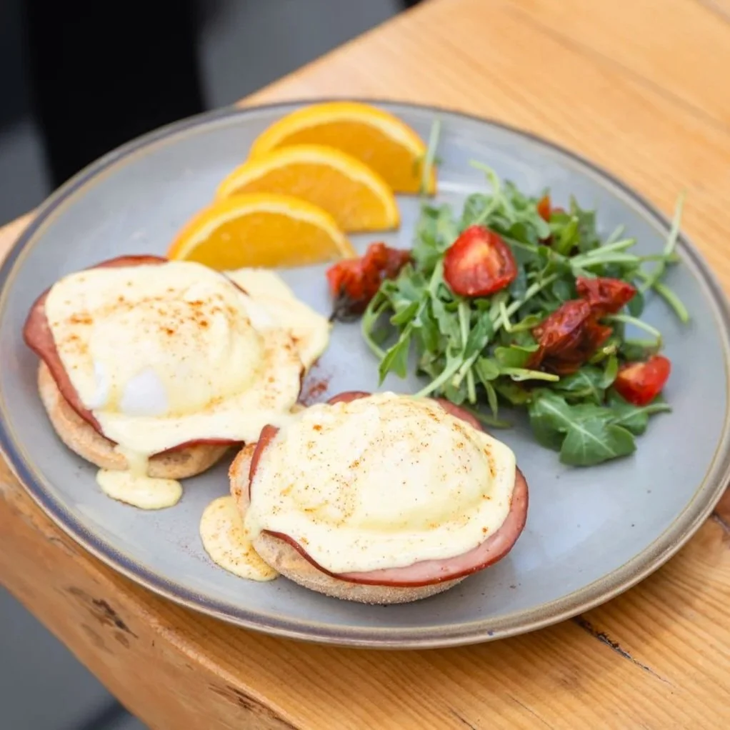 Eggs Benedict with ham, soft poached eggs, and hollandaise sauce served with arugula salad, charred cherry tomatoes, and orange slices.