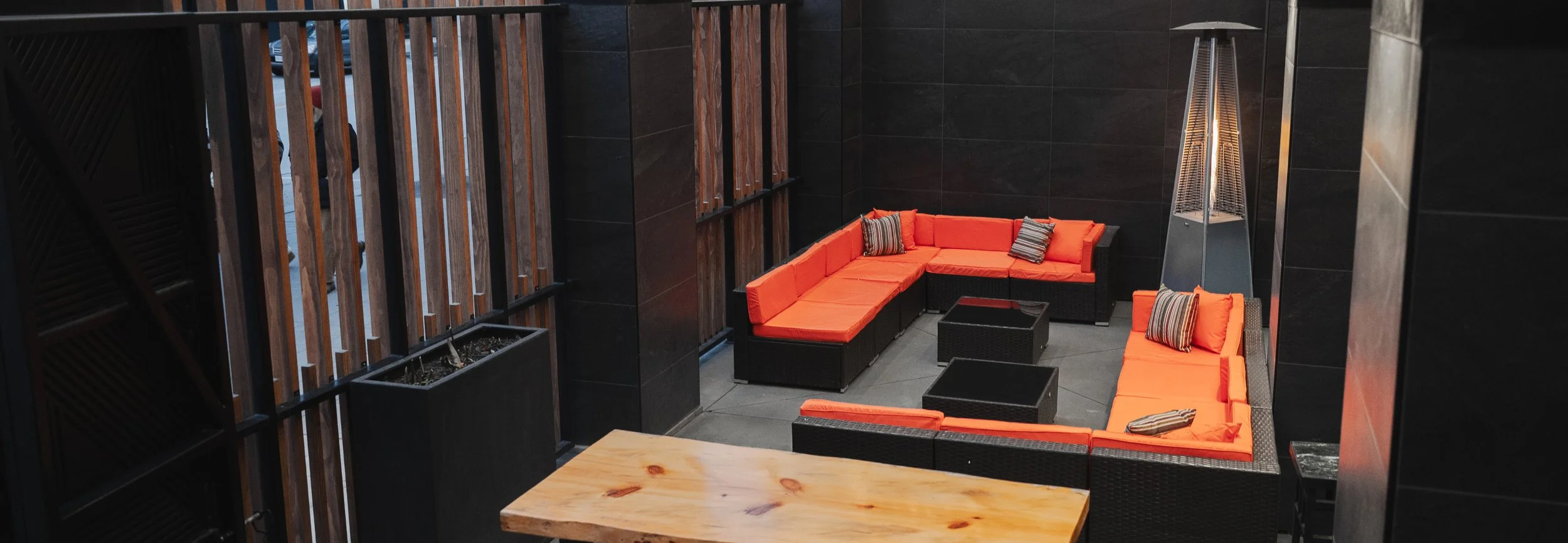 Outdoor enclosed patio featuring black wicker L-shaped sofas with orange cushions, striped accent pillows, a wooden table, and a tall outdoor fire heater.
