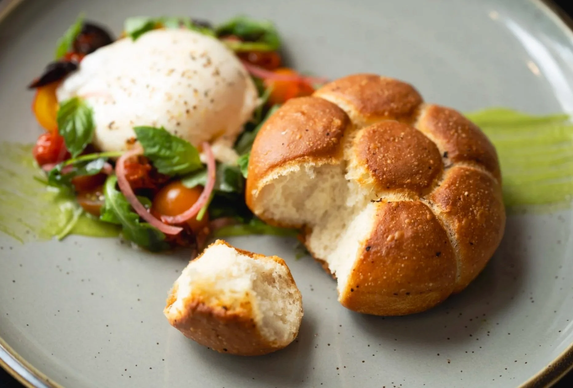 Warm pull-apart focaccia paired with fresh burrata, mixed greens and charred cherry tomatoes, on a dinner plate.