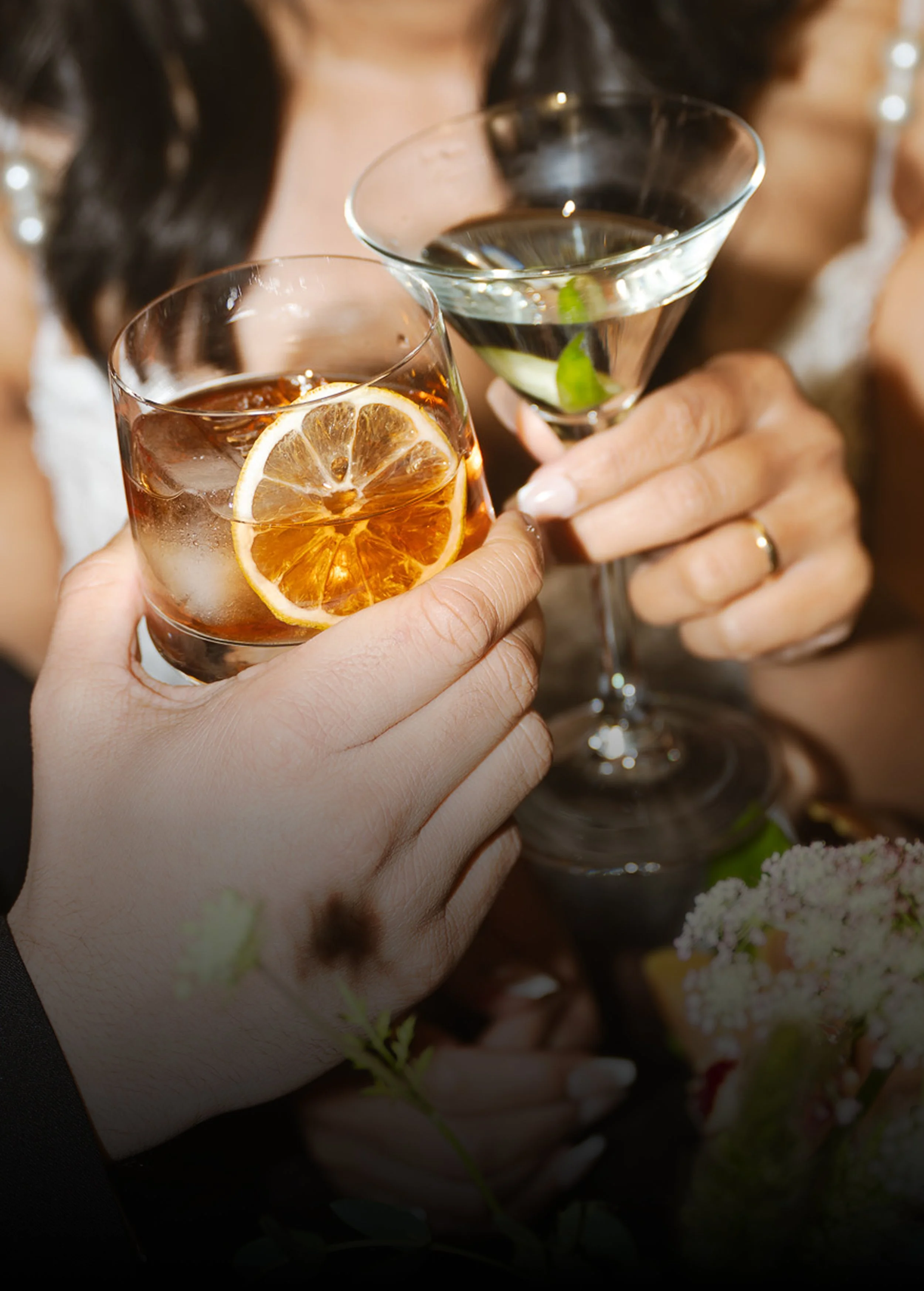 Close-up of a groom holding a whiskey with a lemon garnish and a bride in a white wedding dress holding a martini with a lime garnish while seated together.